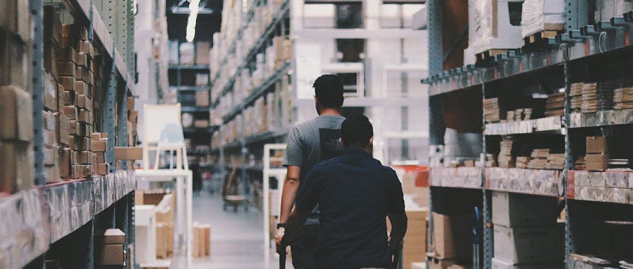 Blick in ein Warenlager in dem zwei Personen mit dem Rücken zum Betrachter ein Paket tragen. Mit der passenden Fufillment-Software automatisierst du deine Prozesse und sorgst für zufriedene Kund:innen!
