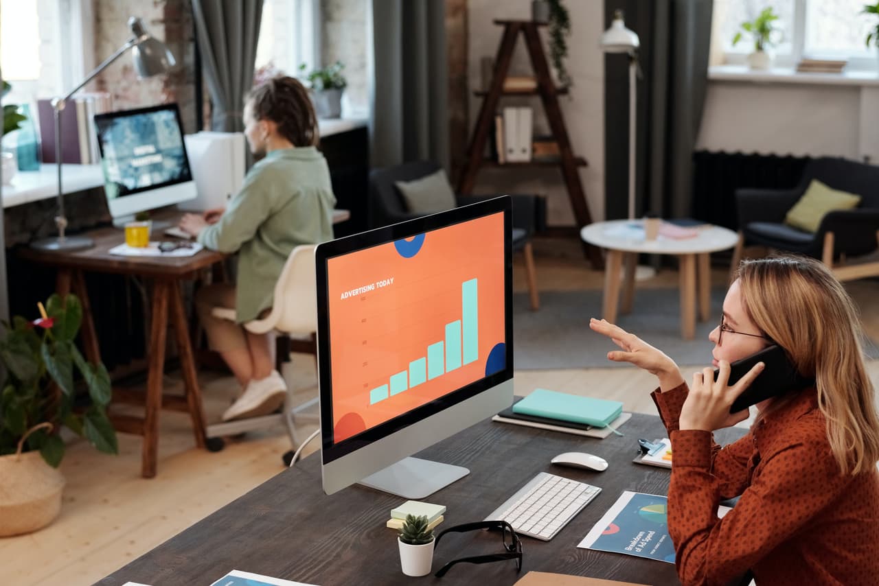 Blick in ein Büro. Im Hintergrund sitzt eine junge Frau an einem Schreibtisch vor einem Bildschirm. Im Vordergrund sitzt eine weitere junge Frau mit einem Handy am Ohr vor einem Schreibtisch mit einem Bildschirm darauf. Auf dem Bildschirm ist ein Balkendiagramm mit ansteigendem Trend zu sehen. Deinen Vertrieb zu automatisieren hat enormes Optimierungspotenzial! Wir zeigen dir in unserem Beitrag, worauf du achten solltest.