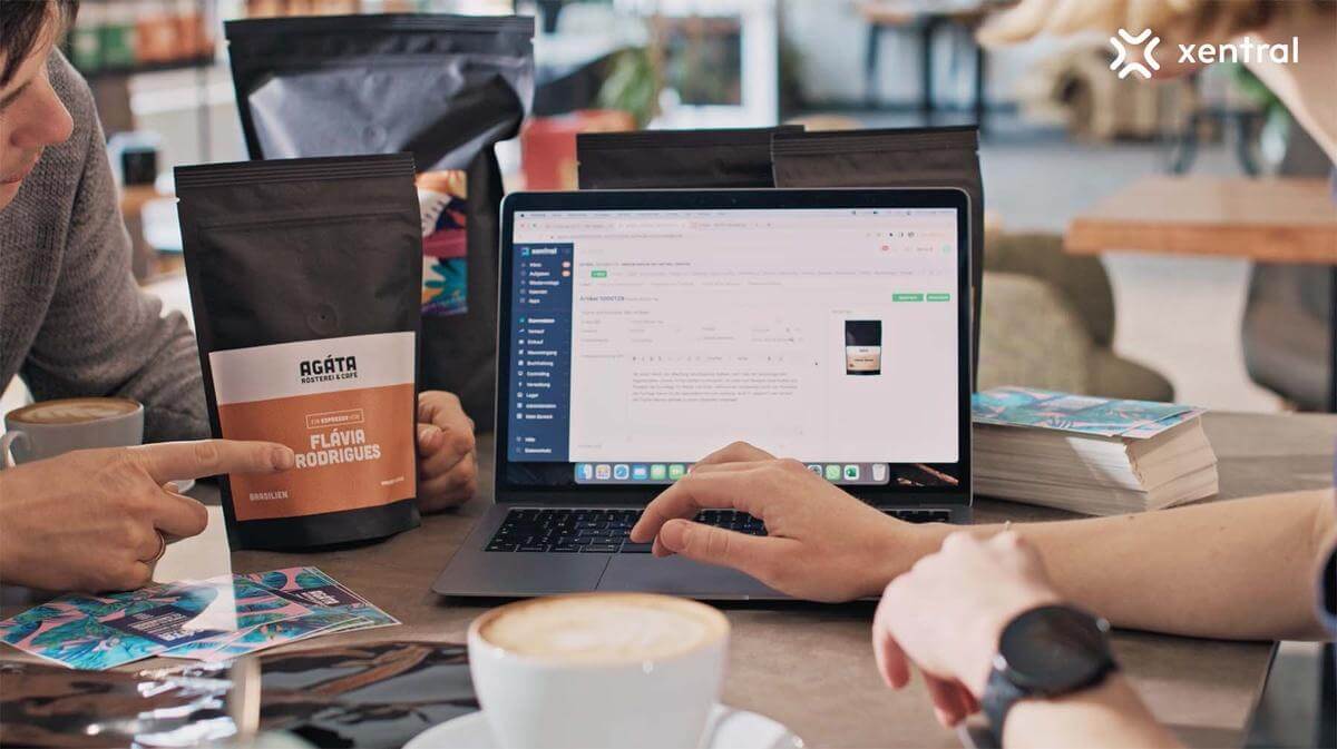 Zwei Personen arbeiten an einem Laptop mit dem Logo von xentral auf dem Bildschirm, neben einer Kaffeetasse und einer AGATA-Kaffeepackung.