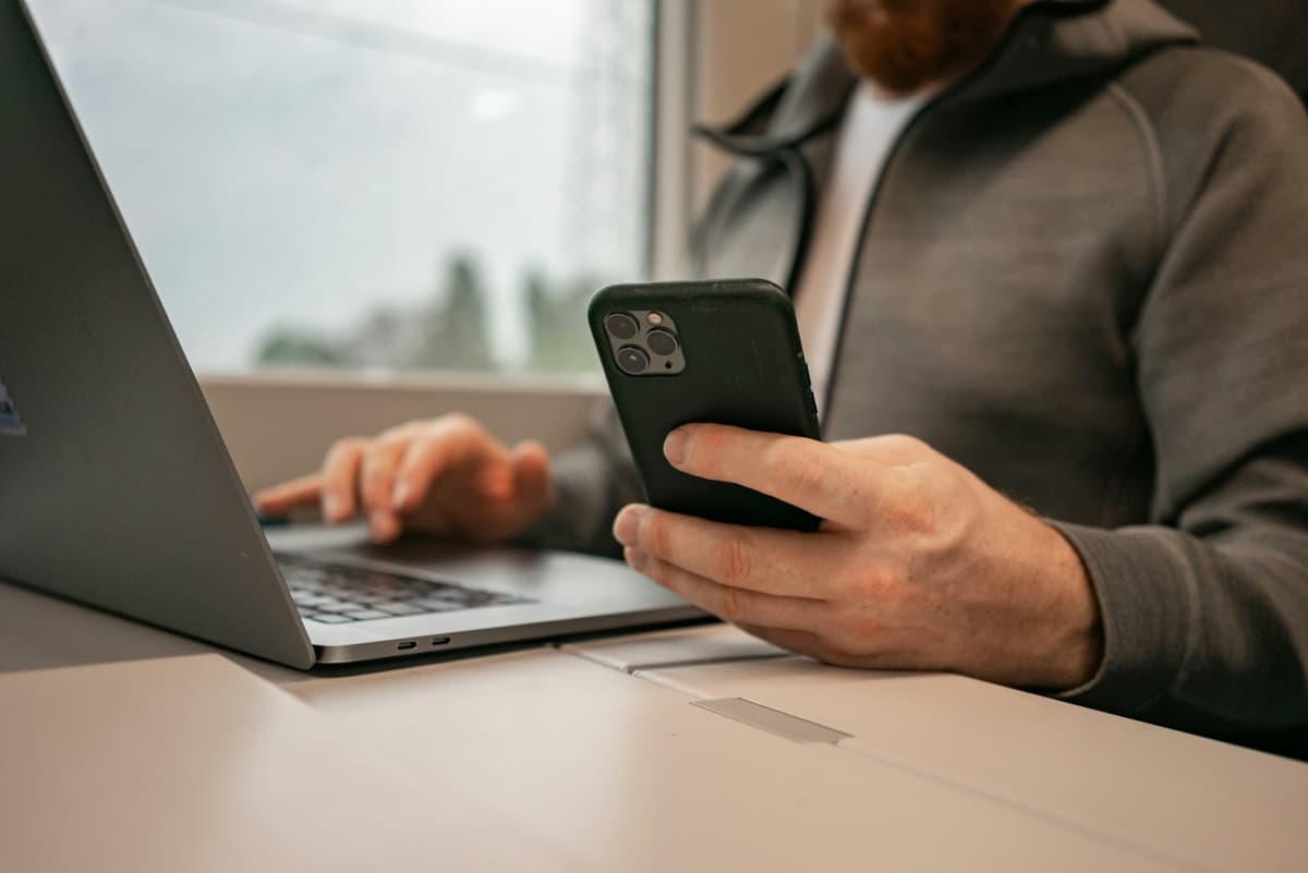 Nahaufnahme eines Mannes, der vor einem Laptop sitzt und in der linken Hand ein iPhone hält, um seine Versandbestätigung zu checken.