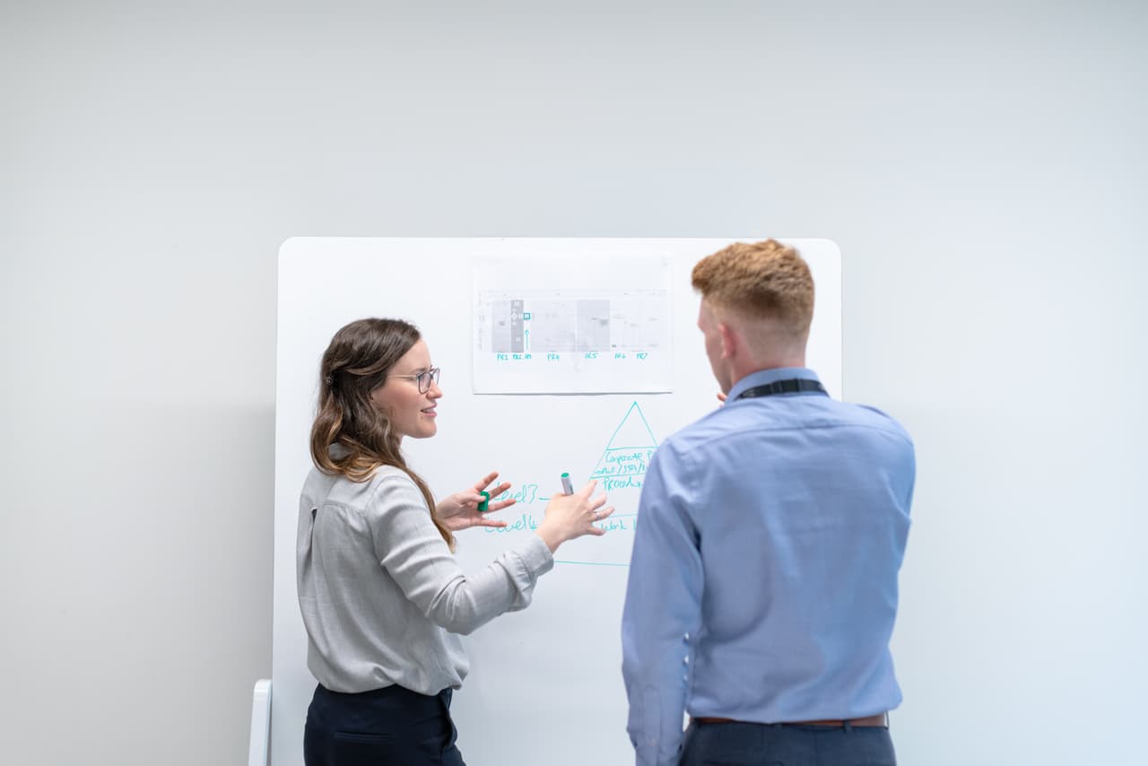 Eine junge Frau und ein junger Mann stehen in einem hellen Raum vor einem Whiteboard vor einer weißen Wand und diskutieren miteinander. Die Entscheidung ERP kaufen oder mieten ist nicht einfach. Wir zeigen dir in unserem Beitrag, worauf du achten musst!