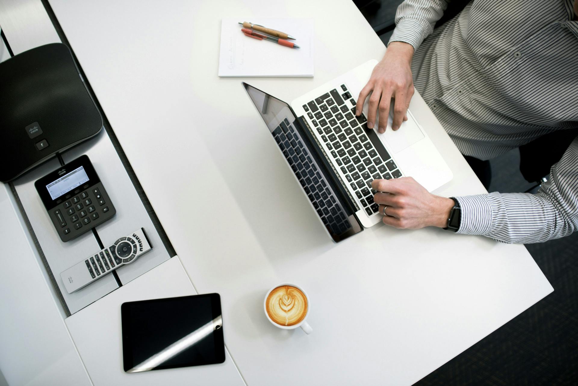 Blick von oben auf einen weißen Bürotisch an dem ein Mann in einem gestreiften Hemd vor einem aufgeklappten Laptop sitzt. Daneben liegt ein Block mit Stiften und es steht eine Kaffeetasse hinter dem Laptop. Wir zeigen dir in diesem Beitrag, was du über E-Rechnungen wissen musst. 