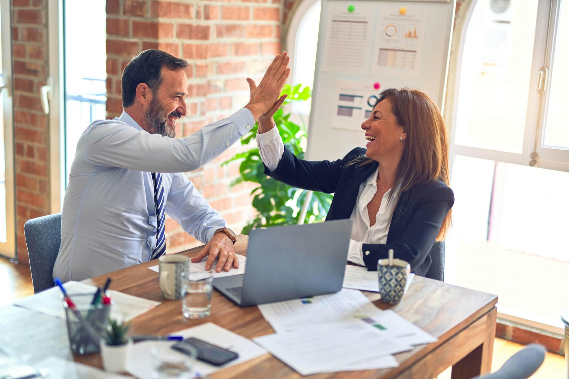 Blick in ein helles Büro an dem ein Mann und eine Frau an einem Holztisch sitzen und sich ein High-Five geben. Du fragst dich, was der Unterschied zwischen ERP & CRM ist? In diesem Beitrag erfährst du mehr!