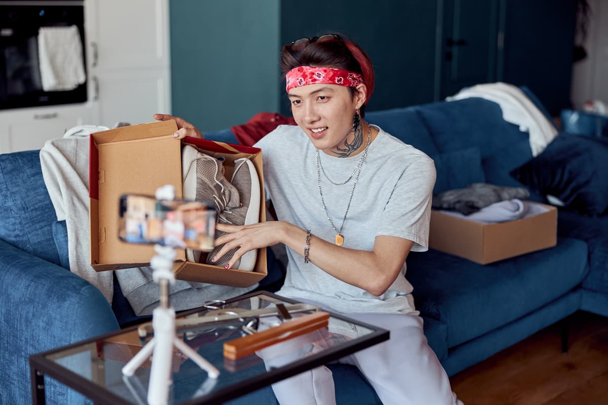 Influencer präsentiert Sneakers in einer Schuhbox während eines TikTok Livestreams. Der Content Creator sitzt auf einem blauen Sofa mit Smartphone auf Stativ und erstellt ein Unboxing-Video für Social Media. Moderner Livestream-Verkauf und digitales Marketing im Home-Setting.