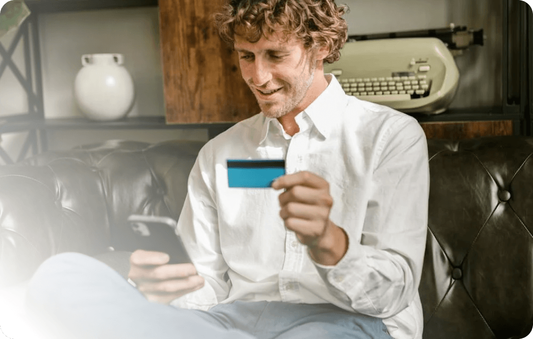 Zufriedene Kunden durch reibungslose Checkout-Prozesse mit Xentral ERP für e-Commerce: Ein lächelnder Mann sitzt auf einer Ledercouch und hält eine Kreditkarte sowie ein Smartphone in den Händen, im Hintergrund ist eine Schreibmaschine in einem Regal zu sehen.