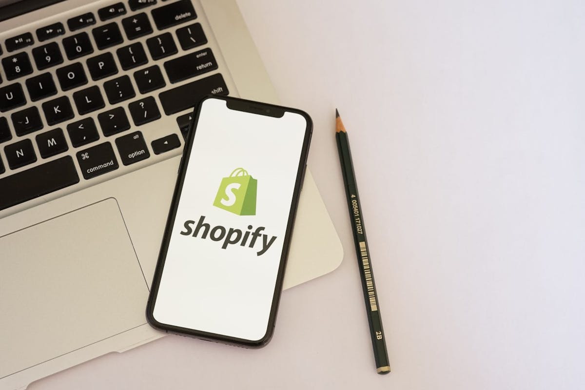Auf einem Laptop liegt ein Handy auf dem das Shopify-Logo zu sehen ist. Warum die Kombination aus Shopify und Xentral die perfekte Lösung ist, erfährst du im Beitrag. 