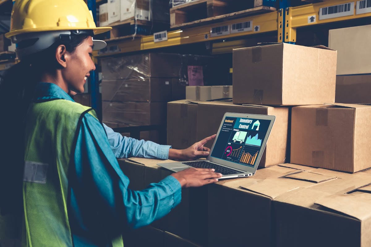 Blick in ein Warenlager in dem verschiedene Pakete gestapelt sind. Eine junge Frau mit einem gelben Sicherheitshelm auf dem Kopf steht vor einem Laptop, der auf den Paketen steht. Eine Online-Warenwirtschaft hilft dir bei der Organisation deiner Warenflüsse!   